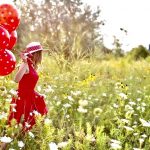 Junge Frau mit rotem Kleid läuft über eine Blumenwiese. In der Hand hält sich viele rote Luftballons